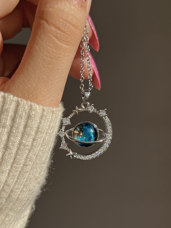 The Infinity Galaxy Necklace, a celestial masterpiece designed to capture the magic of the cosmos. Featuring a luminous deep blue planet orbited by shimmering cubic zirconia stars, this pendant is beautifully framed in a radiant gold-tone setting, perfect for dreamers, stargazers, and lovers of unique jewelry.