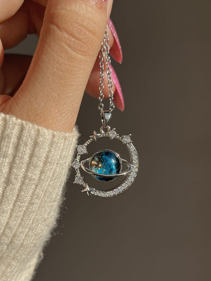 The Infinity Galaxy Necklace, a celestial masterpiece designed to capture the magic of the cosmos. Featuring a luminous deep blue planet orbited by shimmering cubic zirconia stars, this pendant is beautifully framed in a radiant gold-tone setting, perfect for dreamers, stargazers, and lovers of unique jewelry.