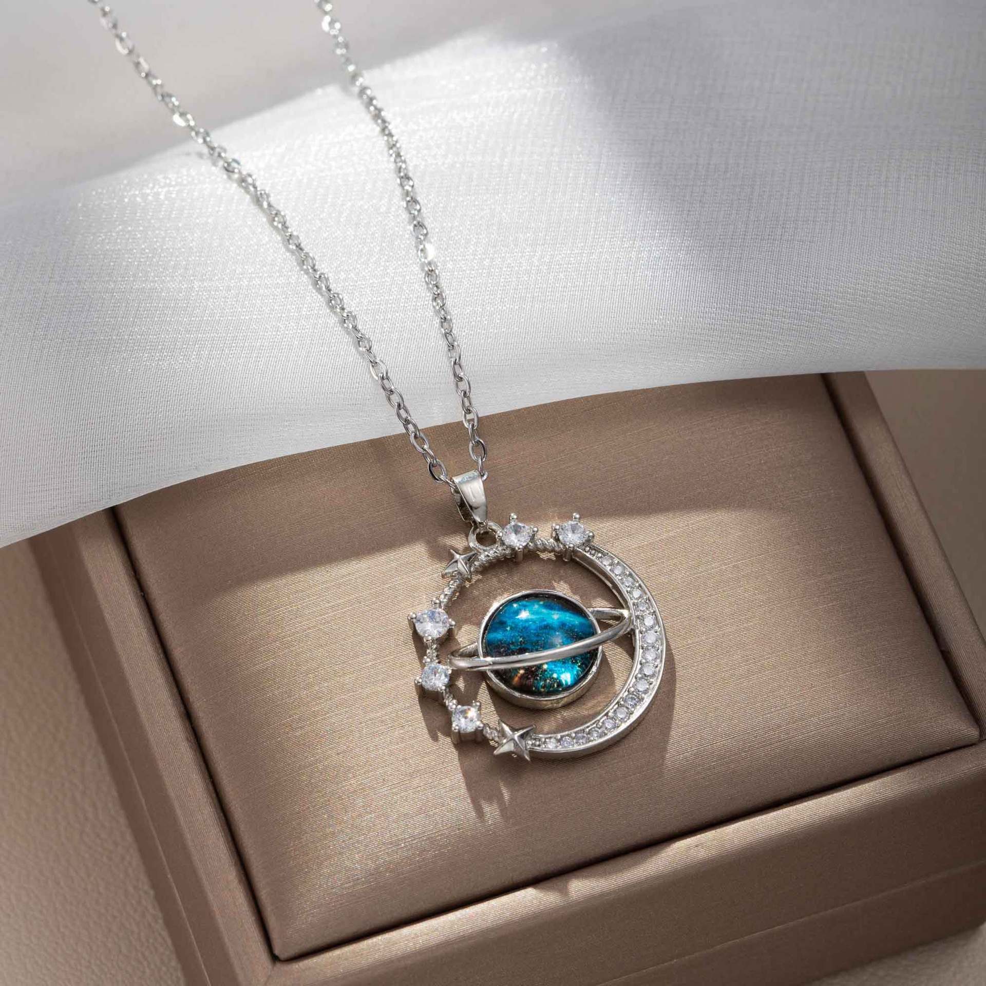 The Infinity Galaxy Necklace, a celestial masterpiece designed to capture the magic of the cosmos. Featuring a luminous deep blue planet orbited by shimmering cubic zirconia stars, this pendant is beautifully framed in a radiant gold-tone setting, perfect for dreamers, stargazers, and lovers of unique jewelry.