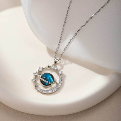The Infinity Galaxy Necklace, a celestial masterpiece designed to capture the magic of the cosmos. Featuring a luminous deep blue planet orbited by shimmering cubic zirconia stars, this pendant is beautifully framed in a radiant gold-tone setting, perfect for dreamers, stargazers, and lovers of unique jewelry.